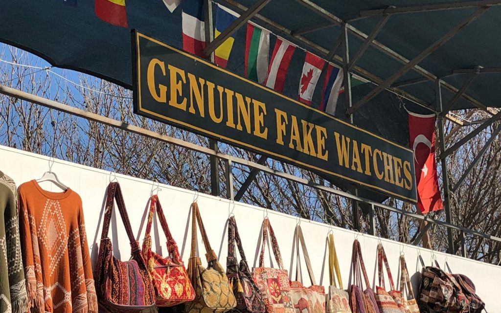 A sign that says genuine fake watches above a bunch of woven bags.