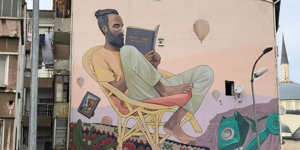 Street art of a man sitting reading a book with hot air balloons in the background and a phone that is off the hook. 