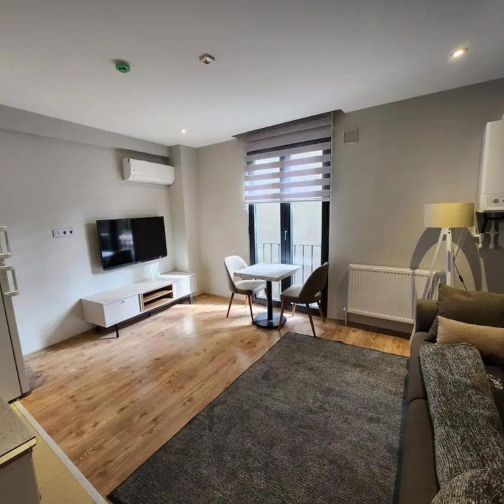 View of the living room where there is a tv, table, kitchenette, air conditioner and a gray couch.