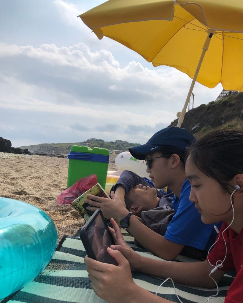 Three people read under a beach umbrella as they lay on the sand.