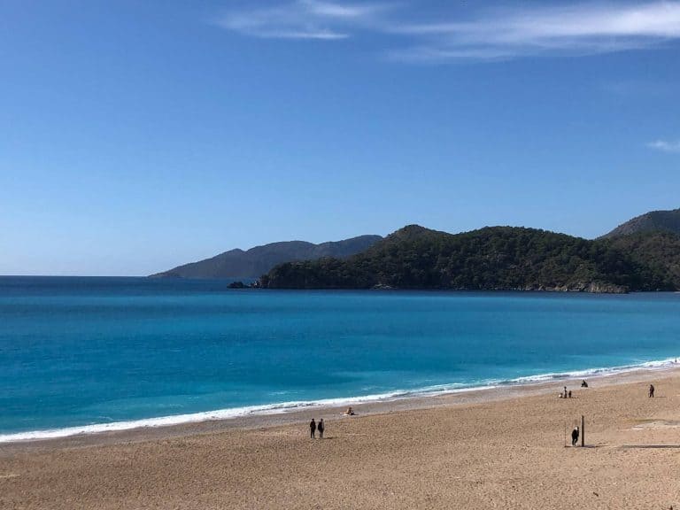A few people enjoy walking down a quiet beach in Fethiye, with a blue sea and mountains in the distance.