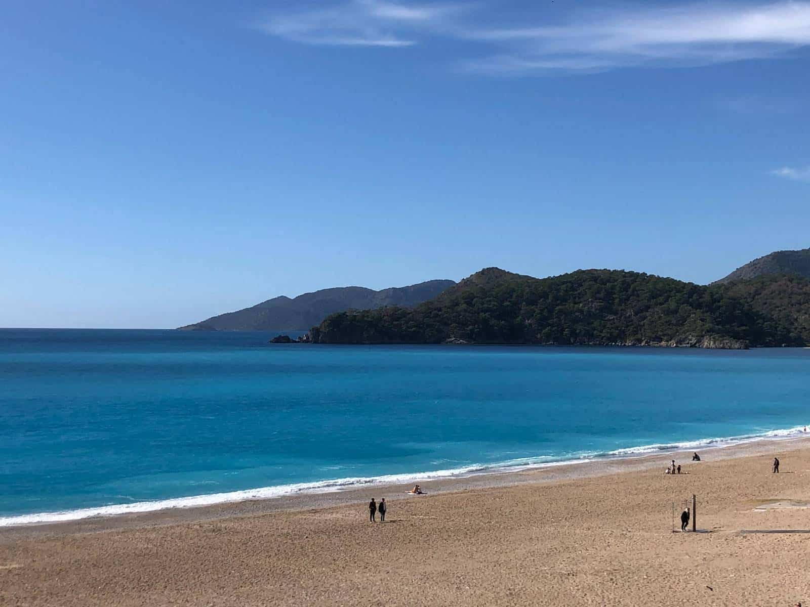 A few people enjoy walking down a quiet beach in Fethiye, with a blue sea and mountains in the distance.
